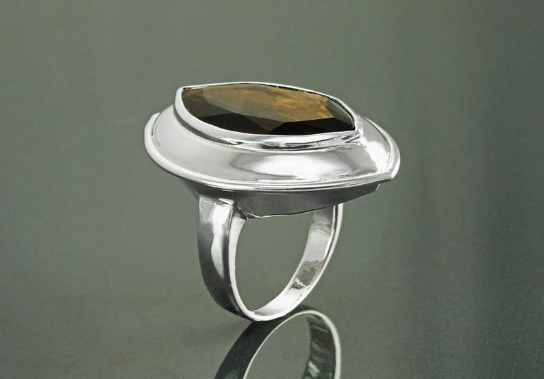 May include: A silver ring with a large, marquise-cut, brown gemstone. The gemstone is set in a wide, oval bezel. The ring has a polished, reflective surface and a simple band. The ring is displayed on a reflective surface.