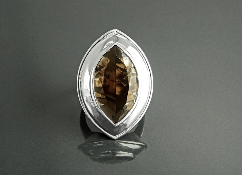 May include: A silver ring with a large, oval-shaped, smoky quartz gemstone. The gemstone is a translucent brown color and is set in a polished silver bezel. The ring is displayed on a reflective surface.