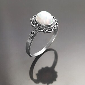 Puede incluir: Un anillo de plata con una gran piedra preciosa de &oacute;palo blanco ovalada. La piedra preciosa est&aacute; engastada en un engaste decorativo de estilo filigrana. La banda del anillo tiene un dise&ntilde;o ornamentado. El anillo se muestra sobre una superficie reflectante.