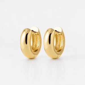 Puede incluir: Un par de pendientes de aro de oro pulido con un perfil grueso y redondeado. Los pendientes presentan una superficie lisa y reflectante con un diseño circular clásico, mostrando un acabado brillante que refleja la luz sobre el metal.