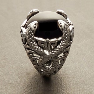 Peut inclure: Une bague en argent avec une pierre précieuse noire sertie dans un motif détaillé de deux serpents entrelacés.
