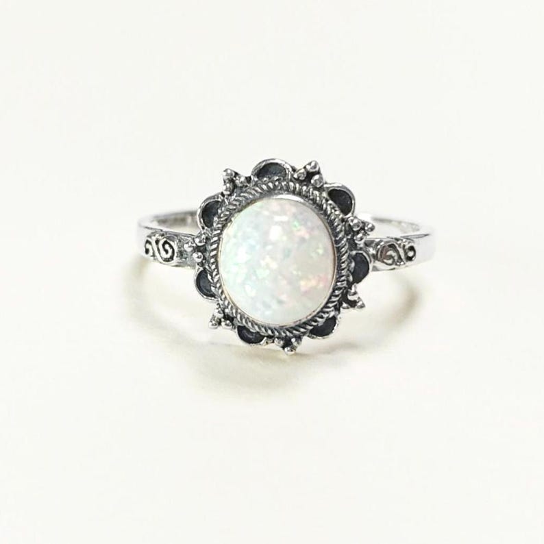 Puede incluir: Un anillo de plata con una gran piedra preciosa ovalada blanca iridiscente. La piedra preciosa est&aacute; engastada en un bisel decorativo de tono oscuro con un dise&ntilde;o floral. La banda tiene detalles ornamentados en los lados, creando una est&eacute;tica vintage o bohemia.