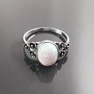 Puede incluir: Anillo de plata con una &oacute;palo blanco ovalado e iridiscente. El anillo tiene un dise&ntilde;o floral a cada lado del &oacute;palo. El anillo est&aacute; sobre una superficie oscura y reflectante.