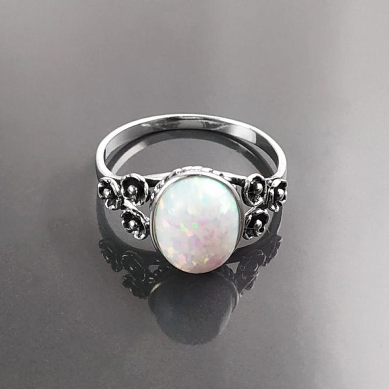 Puede incluir: Anillo de plata con una &oacute;palo blanco ovalado e iridiscente. El anillo tiene un dise&ntilde;o floral a cada lado del &oacute;palo. El anillo est&aacute; sobre una superficie oscura y reflectante.