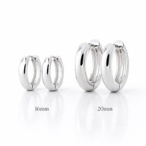 Peut inclure: Deux paires de créoles en argent poli avec fermoirs à charnière. La paire de gauche est étiquetée 16mm et la paire de droite 20mm. Les boucles d'oreilles ont une surface lisse et arrondie avec une finition métallique réfléchissante en deux tailles.
