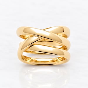 Puede incluir: Un anillo de oro pulido con un diseño de múltiples bandas, donde tres bandas distintas se superponen y se entrecruzan para crear una silueta ancha y estratificada. La superficie metálica es lisa y reflectante, mostrando una estructura continua.