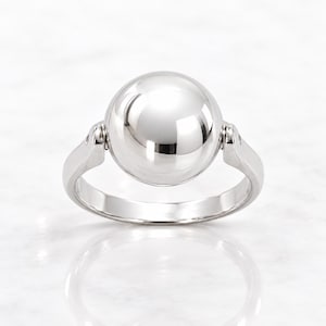 May include: A polished silver-tone ring featuring a large, smooth spherical centerpiece. The sphere is mounted on a hinged mechanism that connects to the band on both sides, allowing the ball to rotate within the frame of the ring setting.