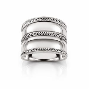 Puede incluir: Un anillo ancho de tono plateado que presenta dos bandas lisas y pulidas, separadas y bordeadas por tres texturas intrincadas de estilo cuerda trenzada. El diseño es simétrico, con los detalles trenzados recorriendo horizontalmente la circunferencia.