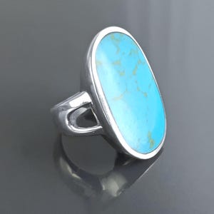 Puede incluir: Un anillo de plata con una gran piedra ovalada de turquesa. La piedra es de un azul vibrante con vetas marrones, engastada en una banda de plata pulida. El anillo tiene un diseño abierto único en los laterales.