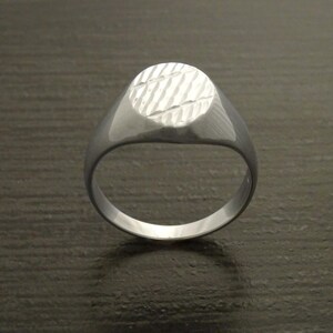 May include: A silver signet ring with a textured oval face. The ring has a smooth, polished band and a detailed, diagonal pattern on the face. The ring is shown on a dark surface.