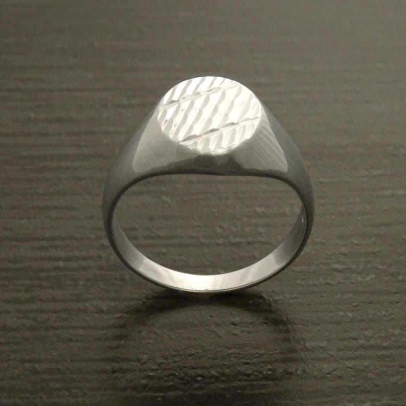 May include: A silver signet ring with a textured oval face. The ring has a smooth, polished band and a detailed, diagonal pattern on the face. The ring is shown on a dark surface.
