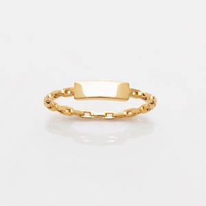Puede incluir: Un anillo de tono dorado que presenta una banda de cadena de eslabones delicados y una placa rectangular lisa y plana centrada en la parte superior. El metal tiene un acabado pulido y reflectante que capta la luz de forma uniforme.