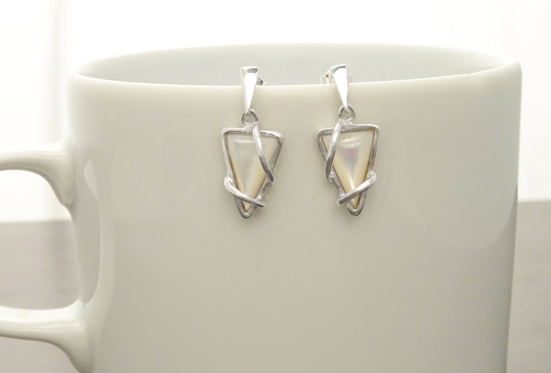 May include: A pair of silver drop earrings featuring a triangular shape with a smooth, iridescent mother-of-pearl inlay. The metal frame wraps around the edges of the triangle, and each earring is suspended from a simple silver post attachment.