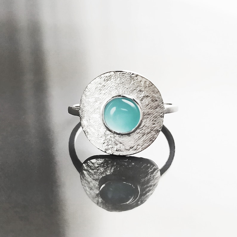 May include: A silver ring with a textured circular band and a light blue gemstone in the center.