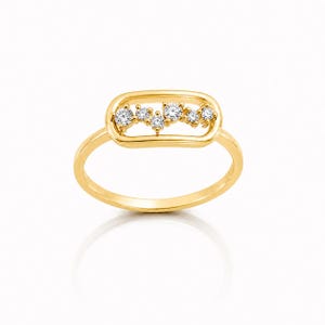 May include: A gold ring featuring a horizontal rectangular frame at the top. Inside the frame, six small, clear, round-cut stones are arranged in a staggered, linear pattern. The band is smooth and polished, creating a continuous gold loop.