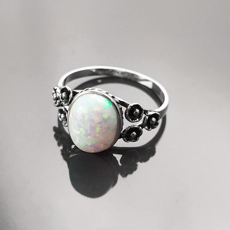 Puede incluir: Anillo de plata con una piedra de &oacute;palo ovalada e iridiscente. El anillo est&aacute; adornado con detalles florales a ambos lados de la gema. El &oacute;palo muestra una mezcla de tonos blancos, verdes y rosas, sobre una superficie reflectante.