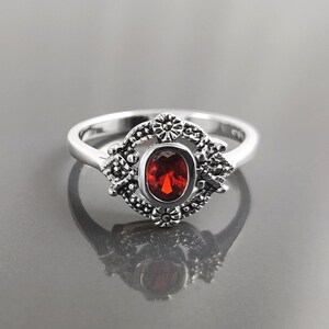 K&ouml;nnte beinhalten: Silberring mit einem ovalen, tiefroten Edelstein. Der Edelstein ist in einem dekorativen, floral inspirierten Rahmen gefasst. Der Ring hat ein glattes, poliertes Band.