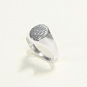 May include: A silver signet ring with an oval face featuring a textured, diagonal striped pattern. The ring has a polished, reflective surface and a classic, minimalist design. The ring is shown on a white background.
