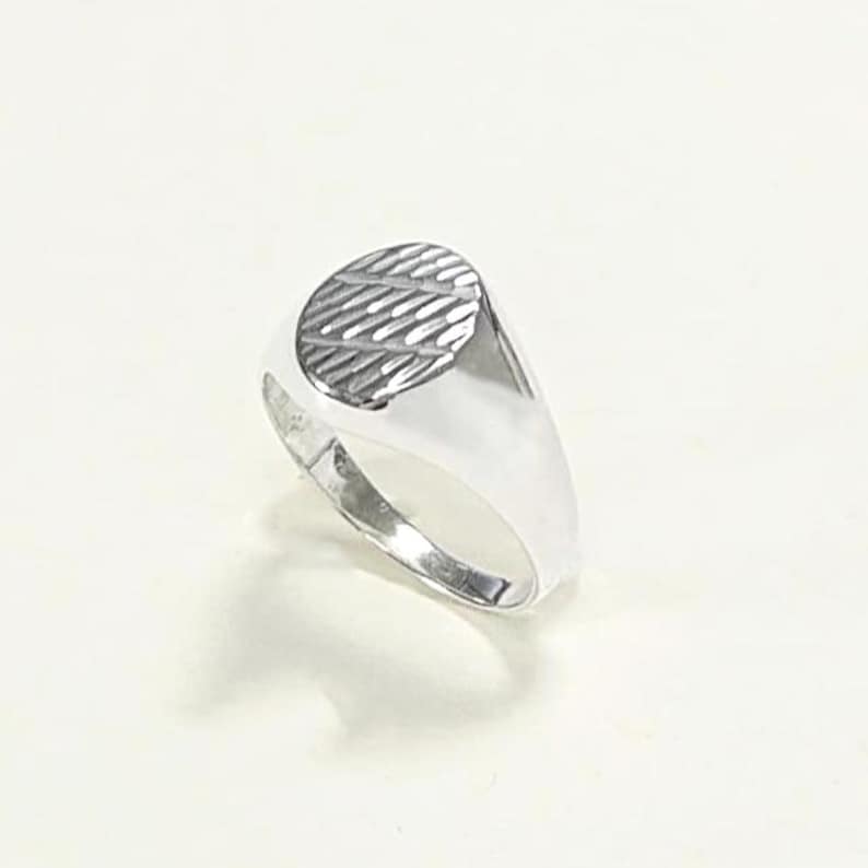 May include: A silver signet ring with an oval face featuring a textured, diagonal striped pattern. The ring has a polished, reflective surface and a classic, minimalist design. The ring is shown on a white background.