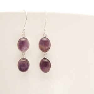May include: A pair of dangle earrings with oval-shaped, purple gemstones set in silver-colored metal. Each earring features two stones, one above the other, connected by a small loop. The earrings hang from a simple hook.