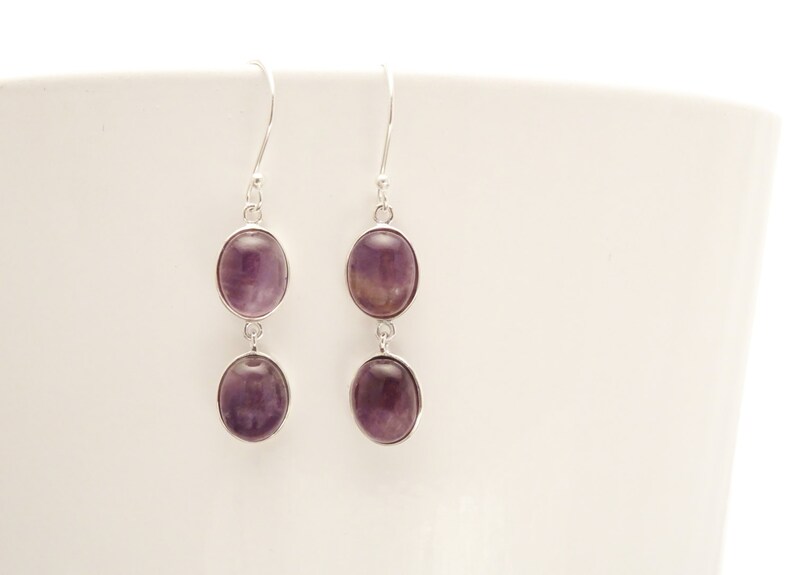 May include: A pair of dangle earrings with oval-shaped, purple gemstones set in silver-colored metal. Each earring features two stones, one above the other, connected by a small loop. The earrings hang from a simple hook.