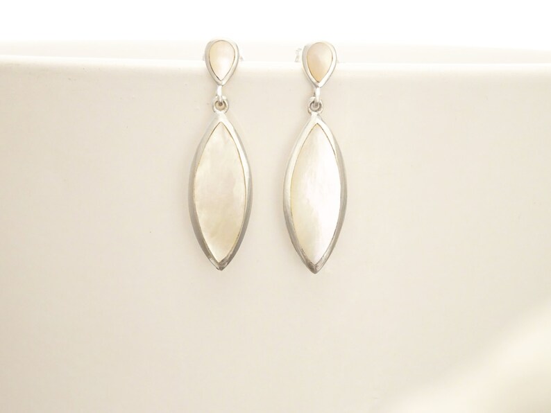 May include: A pair of drop earrings featuring a silver-toned metal frame surrounding an elongated, marquise-shaped white mother-of-pearl inlay. Each earring has a small, teardrop-shaped mother-of-pearl accent at the top, connected by a jump ring.