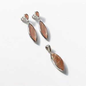 May include: A jewelry set consisting of two drop earrings and one matching pendant. Each piece features a marquise-shaped, iridescent peach-colored stone set in a silver-toned metal frame. The earrings include a small, teardrop-shaped stud top.