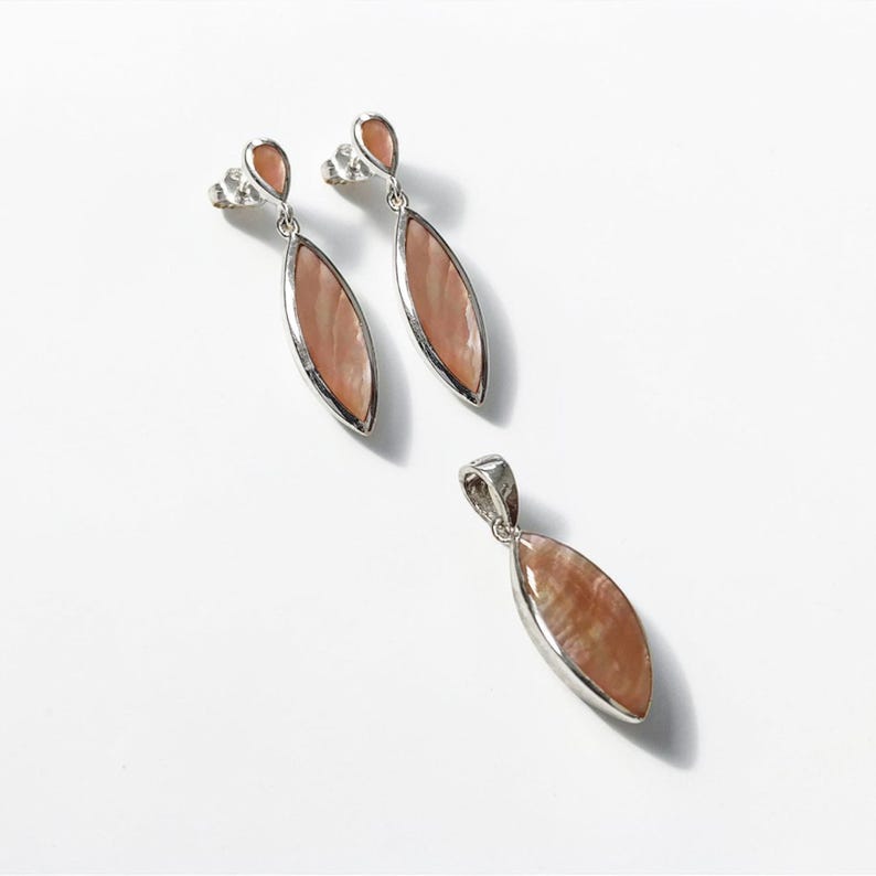 May include: A jewelry set consisting of two drop earrings and one matching pendant. Each piece features a marquise-shaped, iridescent peach-colored stone set in a silver-toned metal frame. The earrings include a small, teardrop-shaped stud top.