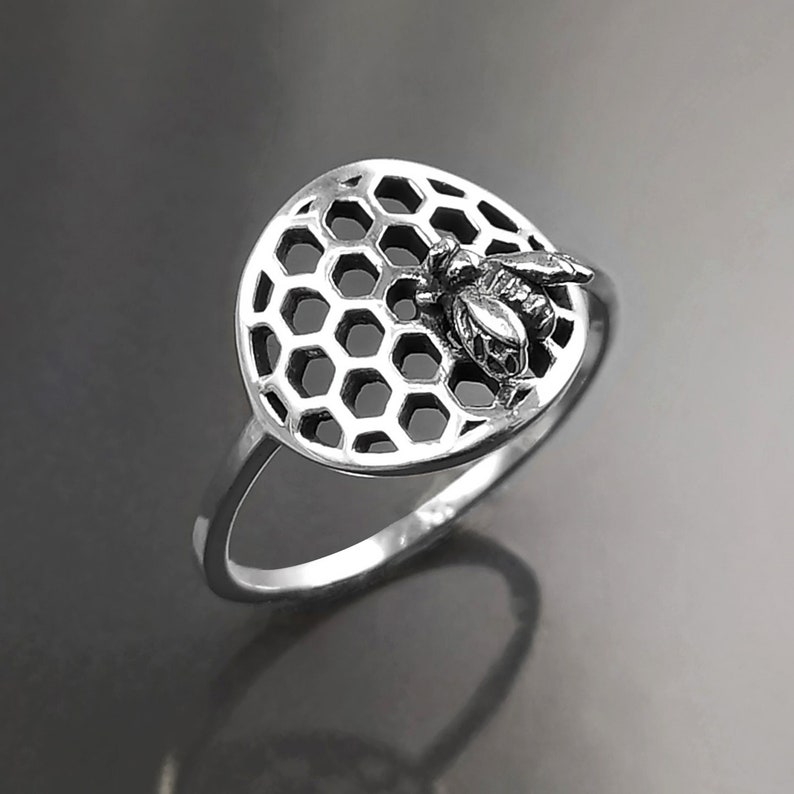 Op de afbeelding: Een zilveren ring met een honingraatontwerp en een bij erop. De ring is een delicaat sieraad met een door de natuur ge&iuml;nspireerd ontwerp.
