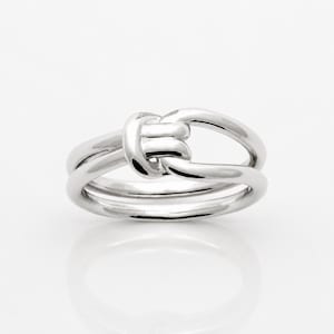 May include: A polished silver-toned ring featuring a central knot design. The band consists of two smooth, interconnected loops that overlap to form a singular, continuous shape. The metal surface is reflective and smooth throughout the entire piece.