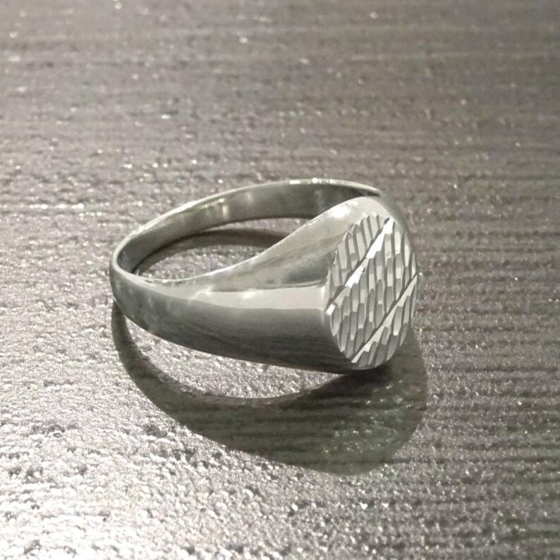 May include: A silver ring with an oval face featuring a textured, diagonal pattern. The ring has a smooth, polished band and a slightly raised face. The ring is shown on a gray surface.