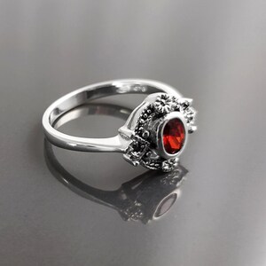 K&ouml;nnte beinhalten: Ein silberner Ring mit einem ovalen, roten Edelstein. Der Ring hat aufwendige Details um den Edelstein herum, mit einem floralen Design. Das Band ist glatt und poliert. Der Ring wird auf einer reflektierenden Oberfl&auml;che gezeigt.