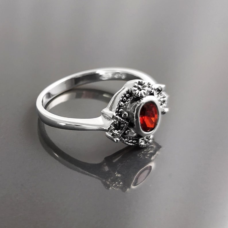 K&ouml;nnte beinhalten: Ein silberner Ring mit einem ovalen, roten Edelstein. Der Ring hat aufwendige Details um den Edelstein herum, mit einem floralen Design. Das Band ist glatt und poliert. Der Ring wird auf einer reflektierenden Oberfl&auml;che gezeigt.