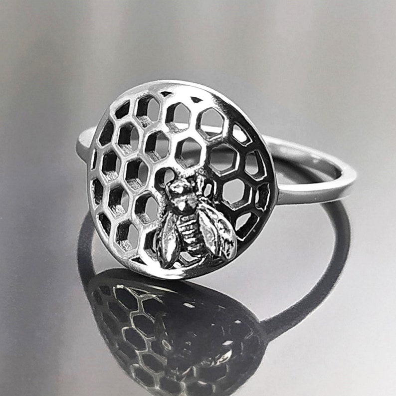 Op de afbeelding: Een zilveren ring met een bij- en honingraatontwerp. De ring is rond en heeft een gedetailleerd honingraatpatroon met een bij in het midden.