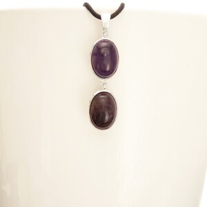 May include: A silver pendant necklace featuring two oval, dark purple gemstones. The gemstones are set in silver bezels and connected by a silver link. The necklace hangs from a black cord.