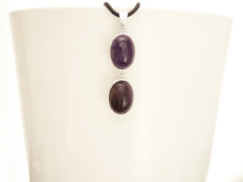 May include: A silver pendant necklace featuring two oval, dark purple gemstones. The gemstones are set in silver bezels and connected by a silver link. The necklace hangs from a black cord.