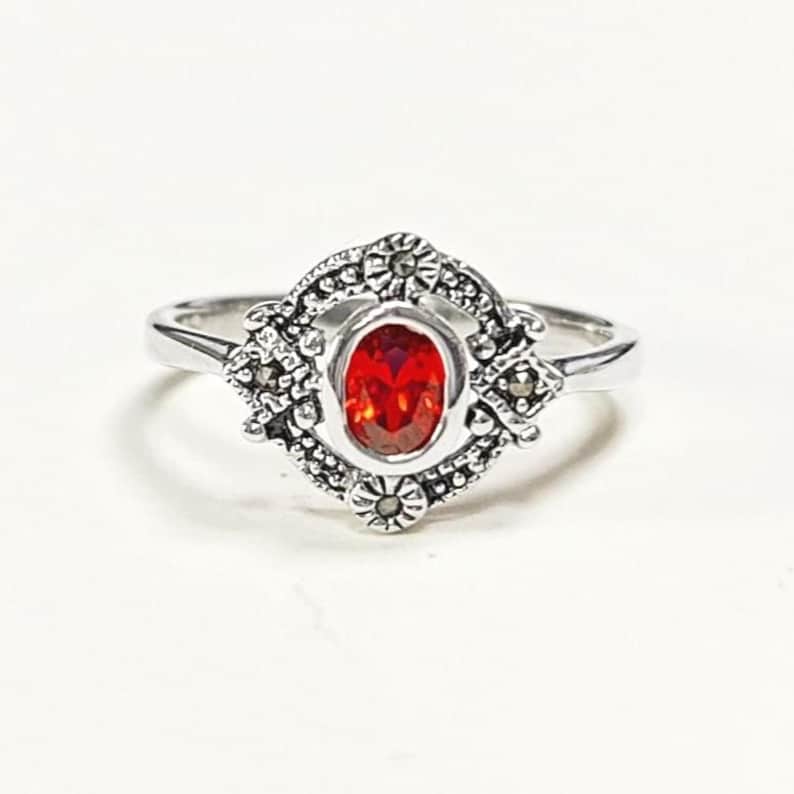 K&ouml;nnte beinhalten: Ein silberner Ring mit einem ovalen, roten Edelstein in der Mitte. Der Edelstein ist von einer dekorativen, floral inspirierten Fassung umgeben. Der Ring hat ein schlankes, poliertes Band und ein elegantes, vintage-inspiriertes Design.
