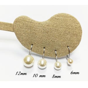 Puede incluir: Exhibición de cuatro pares de pendientes colgantes de perlas, cada uno con una perla de diferente tamaño. Las perlas varían de 6 mm a 12 mm de diámetro, suspendidas de ganchos plateados. Los pendientes se muestran en un expositor de tela beige.