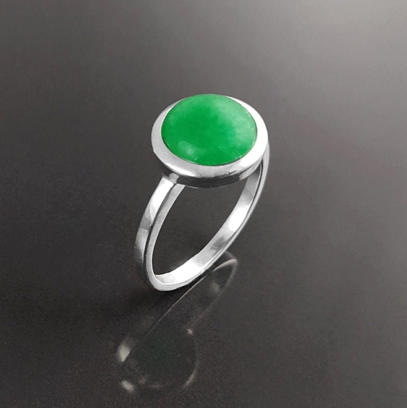 May include: A silver ring featuring a vibrant, round, bright green gemstone. The gemstone is set in a bezel setting, and the ring has a simple, elegant band. The ring is displayed against a dark background.
