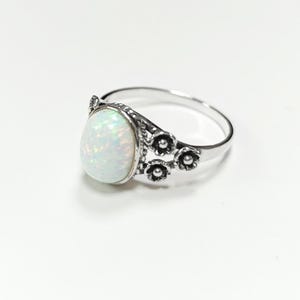 Puede incluir: Un anillo de plata con una piedra preciosa de &oacute;palo ovalada e iridiscente. El anillo tiene un dise&ntilde;o floral en el lateral y una banda pulida. El &oacute;palo muestra un juego de colores, incluyendo blanco, verde y azul. El anillo est&aacute; sobre un fondo blanco.