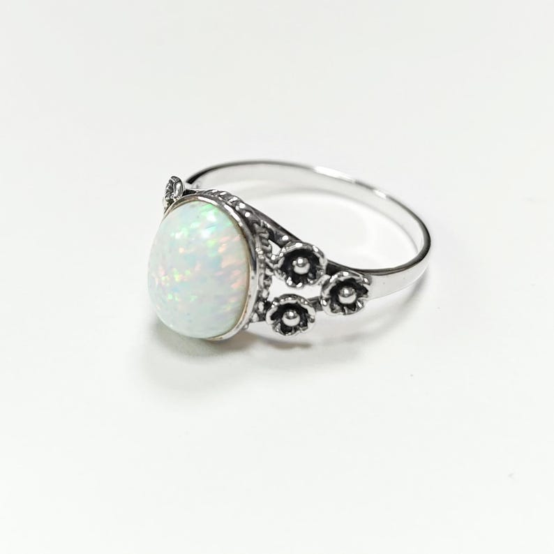 Puede incluir: Un anillo de plata con una piedra preciosa de &oacute;palo ovalada e iridiscente. El anillo tiene un dise&ntilde;o floral en el lateral y una banda pulida. El &oacute;palo muestra un juego de colores, incluyendo blanco, verde y azul. El anillo est&aacute; sobre un fondo blanco.