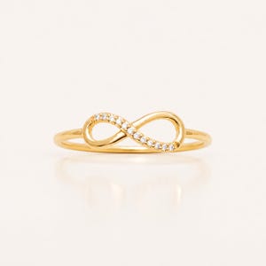May include: A gold ring featuring an infinity symbol centrepiece. The infinity loop is embellished with a series of small, clear, round-cut stones set along the top curve, creating a delicate and polished appearance on a thin, smooth gold band.