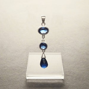 Peut inclure: Un pendentif en argent avec trois pierres précieuses bleues. Le pendentif présente une pierre précieuse en forme de larme en bas, une pierre précieuse ronde au milieu et une pierre précieuse ovale en haut.