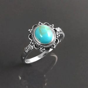 May include: A silver ring with an oval, turquoise gemstone. The stone is set in an elaborate, decorative frame. The ring's band has scroll detailing. The ring is shown against a dark background.