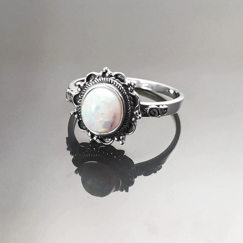 Puede incluir: Un anillo de plata con una piedra preciosa de &oacute;palo ovalada e iridiscente. La piedra preciosa est&aacute; engastada en un bisel detallado y ornamentado. La banda del anillo tiene volutas decorativas. El anillo se muestra sobre una superficie reflectante.
