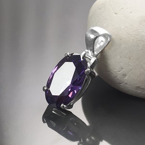 May include: A silver pendant with a large, oval-shaped purple gemstone. The pendant is on a white, smooth surface.
