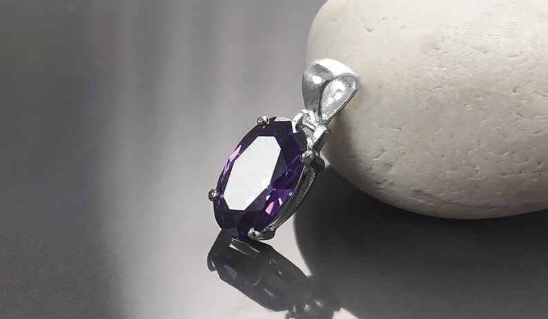 May include: A silver pendant with a large, oval-shaped purple gemstone. The pendant is on a white, smooth surface.