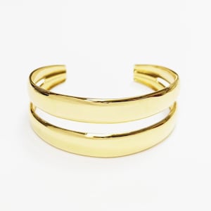 Pode incluir: Uma pulseira de punho dourada com um design de faixa dupla. A pulseira tem uma superfície lisa e polida e um design aberto. A joia está sobre um fundo branco.