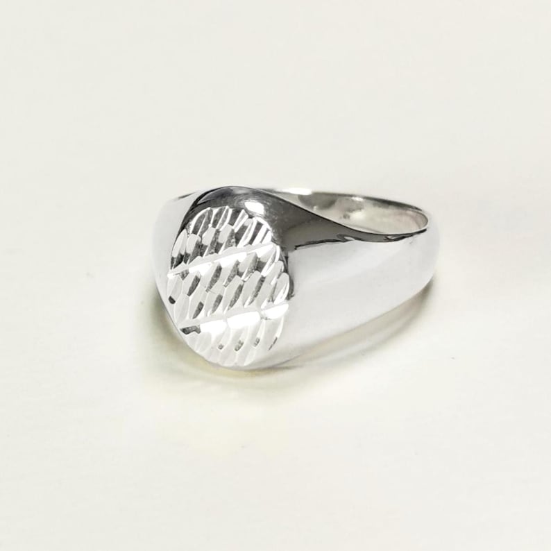 May include: A silver signet ring with a textured oval face. The ring has a polished, rounded band and a detailed, geometric pattern on the face. The ring is shown on a white background.