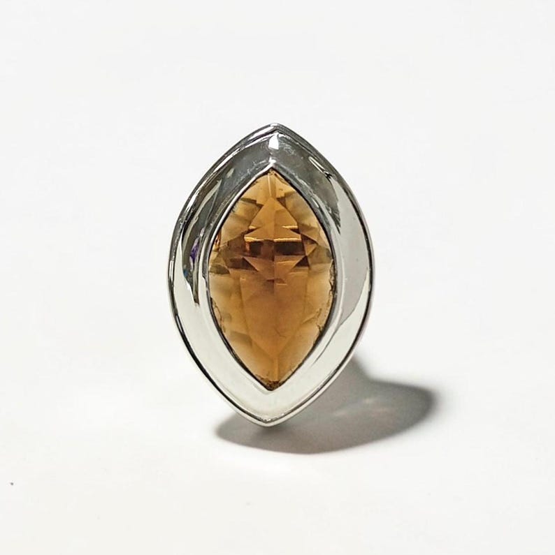 May include: A silver ring featuring a marquise-cut, amber-colored gemstone. The gemstone is set within a polished silver bezel, creating a striking contrast. The ring's design is elegant and eye-catching, suitable for various occasions.